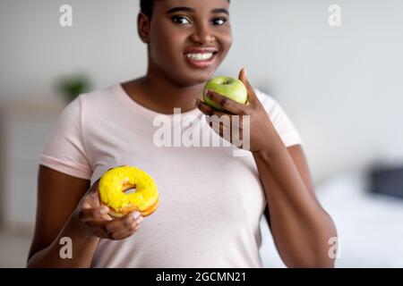 Plump Afro femme choisir entre la pomme et le beignet, choisir des fruits, préférant une alimentation plus saine pour la perte de poids Banque D'Images