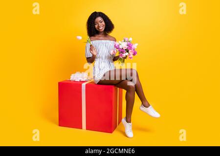 Portrait d'une jolie fille gaie assise sur une grande boîte tenant dans les mains fleurs fraîches isolées sur fond jaune vif Banque D'Images