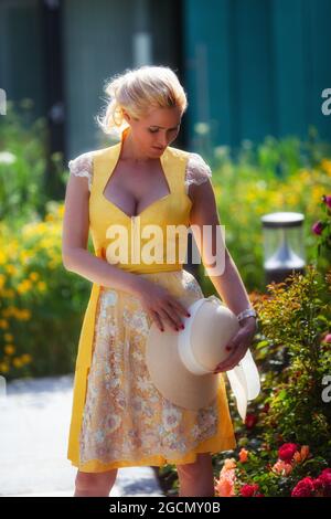 Femme blonde en Dirndl jaune devant la mer des fleurs, avec chapeau de soleil en main. Banque D'Images