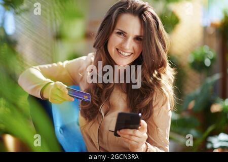 Accueil écologique. Portrait de femme d'intérieur d'âge moyen souriant avec de longs cheveux ondulés avec carte de crédit faisant des achats en ligne sur le site Web d'e-commerce à la maison moderne Banque D'Images