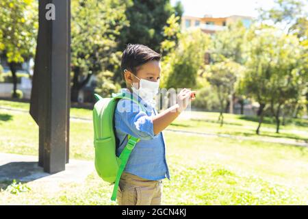 Petit garçon d'âge préscolaire avec masque et sac à dos regardant attentivement une petite branche dans un parc Banque D'Images