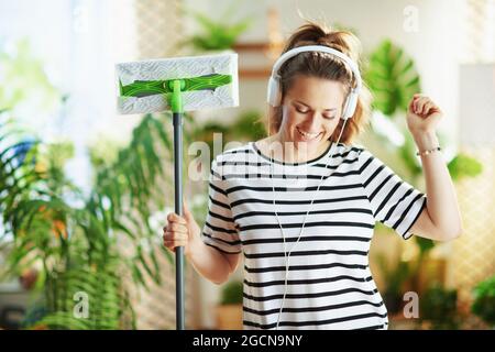 bonne jeune femme en chemise rayée avec mop et casque faisant le travail de ménage dans la maison moderne en journée ensoleillée. Banque D'Images