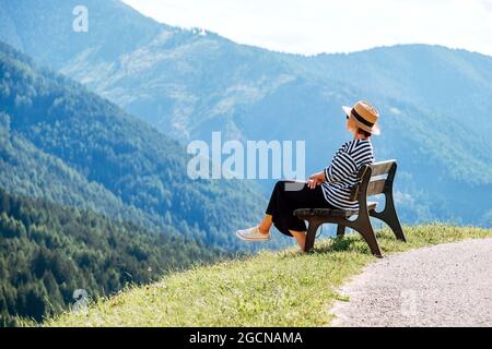 Jeune femme habillée à la mode avec chapeau de paille, assise sur un banc, avec vue sur le village de Santa Maddalena et superbes pics pittoresques des Alpes Dolomites
