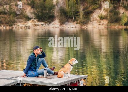 Homme maître de chien et son ami beagle assis sur la jetée en bois sur le lac de montagne et appréciant le paysage pendant leur marche dans les mers d'automne Banque D'Images