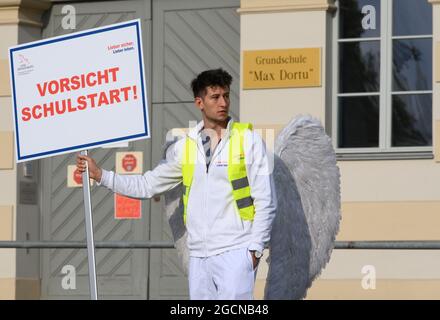 Potsdam, Allemagne. 09e août 2021. Un jeune homme costumé comme un ange de la campagne de sécurité routière se trouve sur la route devant l'école primaire Max-Dortu avec le panneau 'Beware le début de l'école'. Dans les quarante premiers jours après le début de l'école après les vacances d'été, la police veut augmenter les contrôles de la circulation aux heures de priorité. Dans le même temps, la campagne de sécurité routière "Lieber sicher. Lieber leben.' a été lancé pour promouvoir la prise en considération parmi les cyclistes et les automobilistes et pour éduquer les enfants et les parents sur la sécurité routière. Credit: Soeren Stache/dpa-Zentralbild/dpa/Alay Live News Banque D'Images