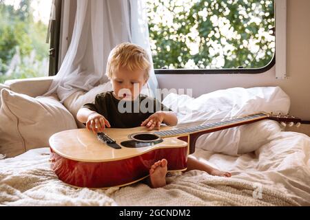 Mignon garçon jouer guitare assis sur le lit dans remorque rv. Adorable petit enfant se divertir en camping-car Banque D'Images