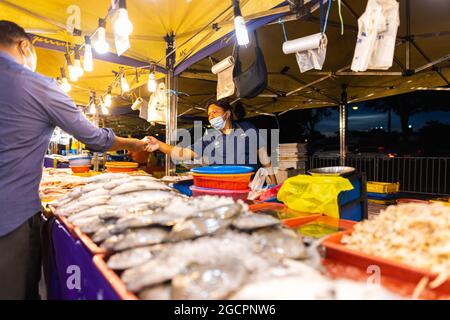 Marché nocturne de la cuisine de rue à Putrajaya, près de Kuala Lumpur. Vendeur avec masque facial dans un magasin de fruits de mer. Le client paie pour le poisson qu'il achète Banque D'Images