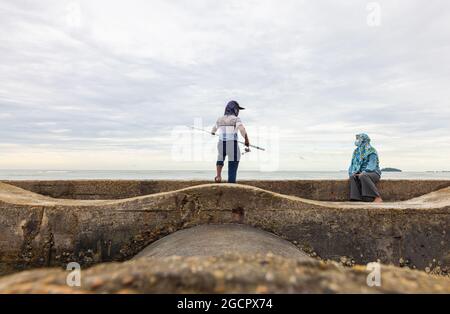 Port Dickson, Malaisie - 15 décembre 2020 : un jeune homme et sa femme pêchent sur un brise-lames sur la plage. Le pêcheur à la ligne jette sa canne à pêche ou son ange dans le Banque D'Images