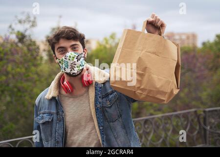 Un adorable gars avec masque facial et écouteurs abaissés montrant un sac à emporter à l'extérieur. Espace pour le texte sur le sac. Banque D'Images