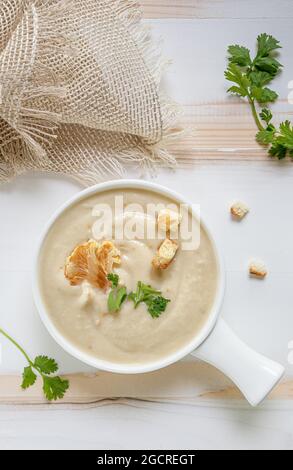 Soupe à la crème de chou-fleur blanc fraîchement préparée avec des herbes vertes fraîches et des croûtons dans un bol en céramique blanche sur une table en bois blanche et beige. Serviette en lin et Banque D'Images