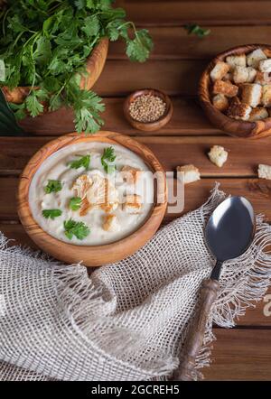 Soupe à la crème de chou-fleur fraîchement préparée avec des herbes hachées et des croûtons sur le dessus dans un bol en bois sur une table en bois brun. Serviette en lin, cuillère, graines de moutarde et Banque D'Images