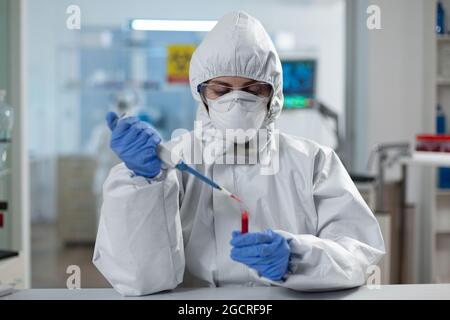 Scientifique femme analysant du sang à l'aide de micropipettes médicales pendant le développement de vaccins covid19 travaillant dans un laboratoire hospitalier microbiologique. Le chercheur porte une combinaison d'epi au cours d'une expérience clinique Banque D'Images