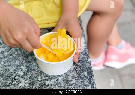 Gros plan d'une fille de 5 ans de race blanche vêtuée d'été et mangeant de la glace jaune à la mangue dans une tasse. Vu en Allemagne en juin Banque D'Images