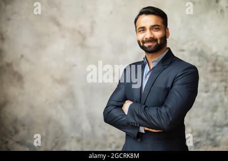 Un jeune homme d'affaires indien d'âge moyen souriant et qualifié en costume formel regarde la caméra, un homme d'affaires arabe à barbe plein d'espoir se tient au bureau avec les bras pliés sur fond gris Banque D'Images