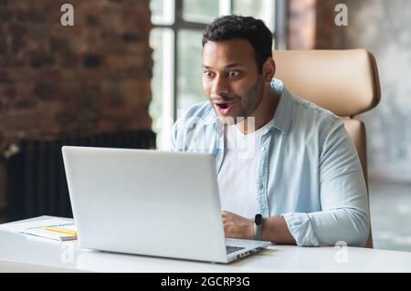 Un homme indien aux cheveux sombres, excité par une chemise décontractée, regardant un ordinateur portable, un entrepreneur indien du Moyen-Orient heureux et choqué a reçu une bonne affaire, e-mails avec une offre d'emploi Banque D'Images