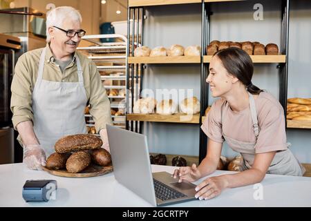 Femme heureuse avec ordinateur portable regardant pile de pains frais sur bois tenu par boulanger mature Banque D'Images