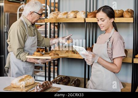 Femme boulangère utilisant une tablette tandis que sa collègue adulte met des petits pains frais en exposition Banque D'Images