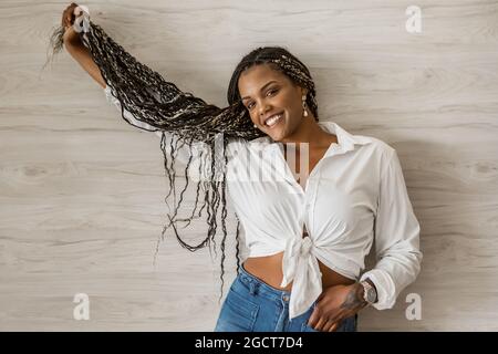 Portrait d'une femme adulte afro-américaine joyeuse tenant et étirant ses cheveux longs. Femme adulte avec des tresses noires et jaunes posant sur un mur ba Banque D'Images