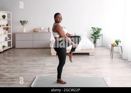 Femme noire en surpoids étirant la jambe, faisant des exercices de flexibilité sur le tapis de yoga, menant le mode de vie actif pendant le covid Banque D'Images