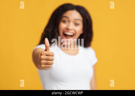 Bonne promotion. Femme noire excitée qui fait un geste du pouce vers le haut à la caméra tout en posant sur fond jaune, mise au point sélective Banque D'Images
