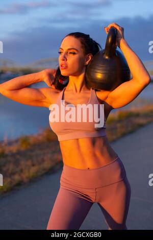 Jeune femme tenant la kettlebell sur l'épaule au bord de la rivière au coucher du soleil doré Banque D'Images