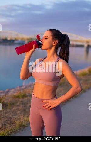 Une jeune femme assoiffée de vêtements de sport boit un rafraîchissement à la bouteille au bord de la rivière Banque D'Images