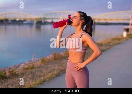 Une jeune femme assoiffée de vêtements de sport boit un rafraîchissement à la bouteille au bord de la rivière Banque D'Images