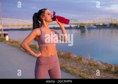 Une jeune femme assoiffée de vêtements de sport boit un rafraîchissement à la bouteille au bord de la rivière Banque D'Images