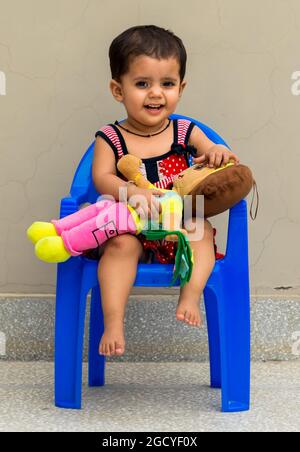 portrait d'une petite fille indienne mignonne assise sur une chaise bleue et jouant avec une poupée de dora. Banque D'Images