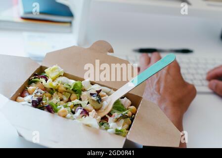 gros plan d'une salade de pois chiches dans un contenant en papier, à côté d'un jeune caucasien assis à son bureau Banque D'Images