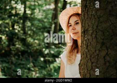 Portrait d'une jeune fille dans la forêt. Elle sort de derrière un tronc d'arbre, une femme dans un chapeau de paille Banque D'Images