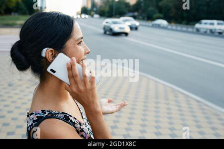 Une femme adulte atteinte d'une déficience auditive utilise une prothèse auditive dans la vie quotidienne, parlant au téléphone dans une ville urbaine en plein air. Solutions auditives Banque D'Images