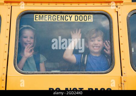 Drôle heureux souriant garçon et fille enfants élèves regardant hors de l'école jaune fenêtre de bus. Agitant de dire Au revoir aux parents avant le début de la journée d'école. CLAS Banque D'Images