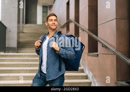 Photo moyenne de jeune homme joyeux et charmant de service de livraison de nourriture avec grand sac à dos thermique posé dans les escaliers du bâtiment de bureau Banque D'Images