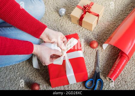 Préparation du papier cadeau des fêtes de Noël, papier d'emballage rouge et boîtes-cadeaux. Femme préparant des cadeaux à la maison. Femme assise sur le sol, cadeau d'emballage b Banque D'Images