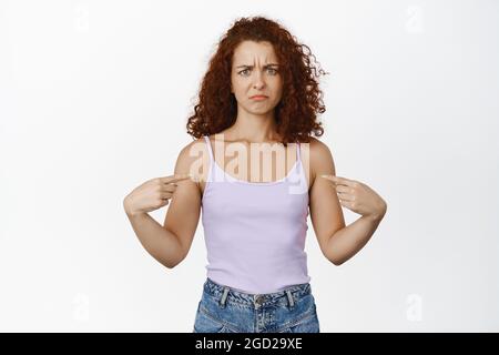 Une femme à tête rouge confuse frognant, pointant ses doigts sur elle-même, offensant ou accusée, debout sur un fond blanc Banque D'Images