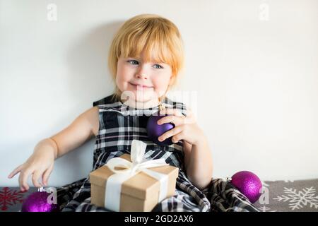 Petite fille souriante avec boîte cadeau de Noël et boules décoratives dans les mains assis sur le lit à la maison dans la robe contre le mur blanc. Préparation des fêtes Banque D'Images