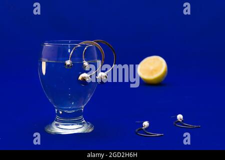 Paires de boucles d'oreilles argentées, un verre d'eau et une tranche de citron sur une surface bleue Banque D'Images