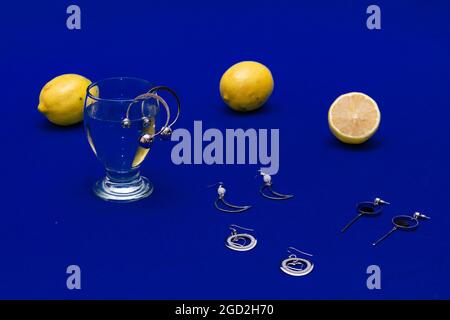 Paires de boucles d'oreilles argentées, un verre d'eau et des tranches de citron sur une surface bleue Banque D'Images