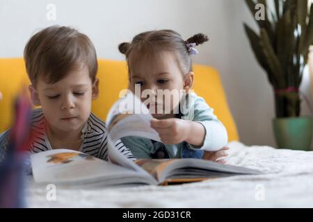 Deux enfants avant-garçonnets lisent un grand livre intéressant de contes de fées sur un lit jaune. Frères et sœurs petits jumeaux lecteur ont plaisir, heureux enfant sur Banque D'Images