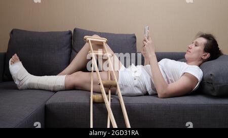 Femme dans un plâtre se trouve sur le canapé à la maison et à l'aide d'un smartphone. Banque D'Images