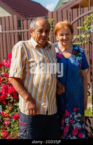 Portrait vertical général d'un couple âgé souriant pendant la journée d'été. Une bonne paire de personnes âgées, mari et femme, est assise sur le jardin fleuri. 60s ou Banque D'Images