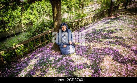 Femme musulmane âgée dans un parc public assis sous un arbre au-dessus d'un lit de fleurs. Elle a l'air heureuse et souriante de regarder l'appareil photo Banque D'Images