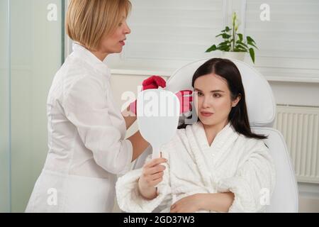 Portrait de la belle femme en peignoir blanc assis sur le lit et de vérifier les résultats du traitement de soin de peau avec cosmetologiste regardant dans le miroir Banque D'Images