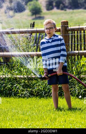 Jeune garçon pulvérisant de l'eau avec un tuyau de jardin dans une cour herbeuse Banque D'Images