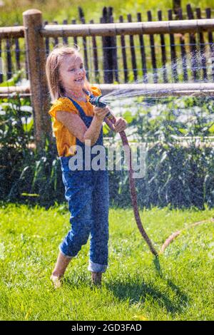 Une jeune fille vaporise de l'eau avec un tuyau de jardin dans une cour herbeuse Banque D'Images