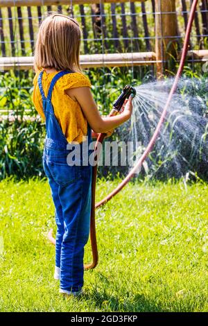 Une jeune fille vaporise de l'eau avec un tuyau de jardin dans une cour herbeuse Banque D'Images
