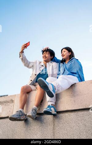 Heureux jeune couple asiatique assis dans la ville posant et prenant le selfie avec leur téléphone. Banque D'Images