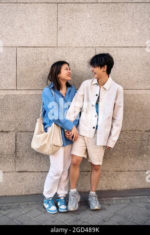 Un jeune couple asiatique se tenant contre un mur gris riant et se regardant l'un l'autre. Banque D'Images