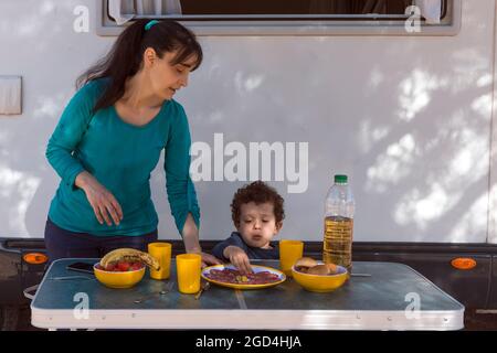 Maman servant de la nourriture à son fils devant la maison du moteur, à la table à l'ombre de l'arbre. Banque D'Images